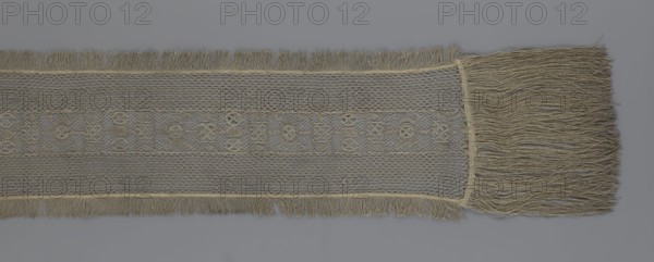 A so-called Saxon lace belt, lace belt accessory for men's clothing. The central part is a continuous belt with a geometric ornament, including a motif of a circle containing an hourglass. The side belts of similar width are permanently connected. The entire belt is trimmed with fringe. 17th/18th century, Poland; lace clothing accessory; height 270 cm, width 25 cm., A long, narrow piece of woven fabric with fringe on both short ends and along one long edge, displayed on a gray background; the fabric shows an open, net-like weave in a beige or tan color with a central decorative band featuring a repeating geometric motif woven in the same beige or tan hue, and a denser woven border separating the decorative band from the netting; a wide tassel-like fringe of loose beige or tan threads extends from one short end.