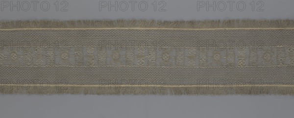 A so-called Saxon lace belt, lace belt as a men's clothing accessory. The central part is a continuous belt with a geometric ornament; among the motifs is a circle containing an hourglass. Side belts of similar width are permanently connected. The entire belt is trimmed with fringe. 17th–18th century, Poland. Materials: lace; dimensions: height 270 cm, width 25 cm., A long horizontal band of woven material with fringed edges runs across the image, showing a central decorative strip flanked by two patterned sections; the central strip contains a repeating motif of interlocking circular and oval shapes set against a textured background, while the adjacent sections display a mesh-like diamond pattern. The band and fringe are predominantly shades of beige and tan with subtle darker brown accents, and the background behind the band is a uniform muted gray.