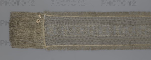 A so-called Saxon lace belt, lace, belts, clothing accessories, men's clothing, clothing, fashion, deposit provenance. The central part is a continuous belt with a geometric ornament; among the motifs is a circle containing an hourglass. The side belts of similar width are permanently connected. The entire belt is trimmed with fringe. 17th/18th century, Poland. Lace, clothing accessories, clothing. Height 270 cm, width 25 cm., A long woven textile strip lies flat against a gray background, showing a central band with a repeating geometric openwork pattern flanked by finer mesh panels and fringed edges; the textile and its fringe appear in shades of tan, beige, and muted gold, with the mesh areas showing slightly darker and lighter variations of those colors, and a small cluster of tan-colored tags or paper pieces rests near one end on top of the fringe.