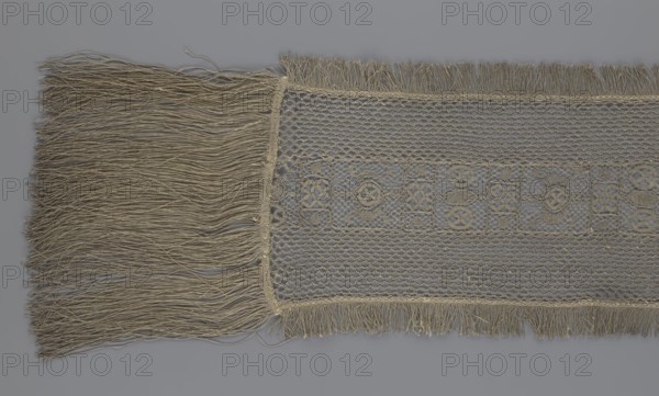 A so-called Saxon lace belt, creator unknown, lace belt for men's clothing, clothing accessory. The central part is a continuous belt with a geometric ornament; among the motifs is a circle containing an hourglass. Side belts of similar width are permanently connected. The entire belt is trimmed with fringe. 17th–18th century, Poland. Materials: lace. Dimensions: height 270 cm, width 25 cm., A section of woven textile with a long fringe on one side and a shorter fringe along the opposite edge, showing a central panel of openwork lace with repeating geometric motifs and a surrounding net-like pattern; the fibers and threads appear in shades of beige and tan against a neutral gray background.