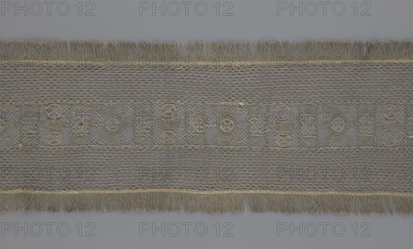 A so-called Saxon lace belt, lace belt and clothing accessory for men's clothing, decorated with a continuous central belt featuring a geometric ornament including a circle containing an hourglass. Side belts of similar width are permanently connected. Entire belt trimmed with fringe. 17th–18th century, Poland. Materials: lace. Dimensions: height 270 cm, width 25 cm., A horizontal band of woven fabric displayed against a gray background, featuring a central decorative strip with an openwork pattern of interlocking shapes in beige and tan tones, flanked above and below by sections of netlike mesh in similar beige and light brown hues, and finished at the top and bottom edges with fringe strands in beige and light tan.