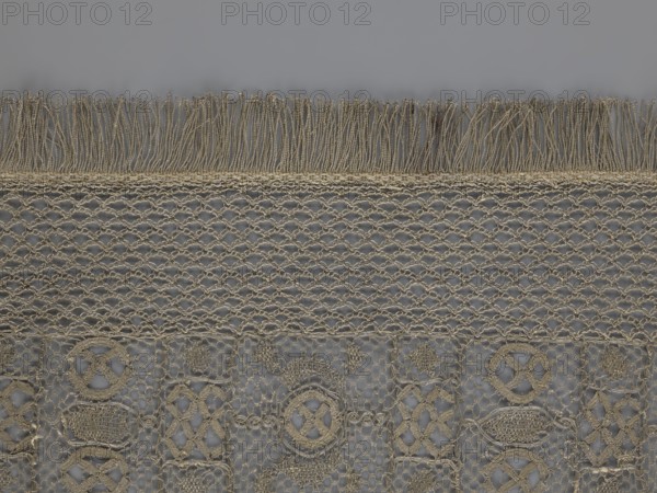 A so-called Saxon lace belt, lace belt as men's clothing accessory. The central part is a continuous belt with a geometric ornament; among the motifs is a circle containing an hourglass. Side belts of similar width are permanently connected. The entire belt is trimmed with fringe. 17th/18th century, Poland. Materials: lace; dimensions: height 270 cm, width 25 cm., A close-up of a textured fabric showing a horizontal band of fringe at the top and an open net-like weave beneath it, followed by a patterned lace section with repeating geometric motifs. The visible colors are beige and gray, with beige thread forming the fringe, netting, and decorative shapes against a gray background.