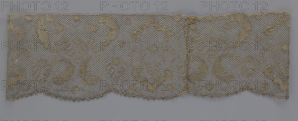 Lace fragment, lace, 18th century, Europe, fabric, width 20 cm, length 120 cm, The image shows a horizontal strip of lace with a scalloped lower edge, featuring a repeating pattern of curved paisley-like motifs and small round shapes set on a hexagonal net ground; the lace appears in shades of beige and gold against a muted gray background, with some areas showing frayed threads and slight irregularities along the top edge.