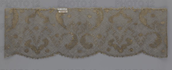 Lace fragment, lace, 18th century, Europe, fabric, width 20 cm, length 120 cm, purchased provenance, A rectangular piece of lace fabric laid flat, showing a net-like background in gray with ornate embroidered motifs in a muted gold; the lace has a scalloped lower edge and a straight upper edge with some fraying, and the gold embroidery forms flowing curved shapes and small filled spots distributed across the gray mesh, with a few loose threads visible along the edges.