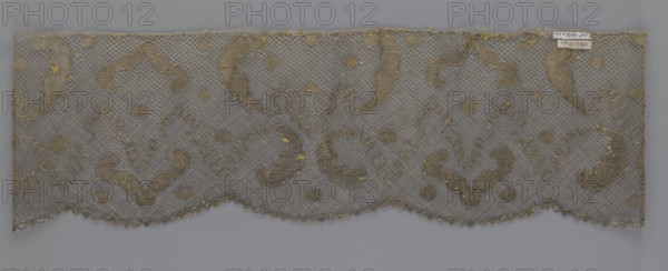 Lace fragment, lace, 18th century, Europe, lace, fabric, width 20 cm, length 120 cm, A rectangular piece of delicate netted fabric with a scalloped lower edge, showing a repeating ornate pattern of curved motifs and small round motifs worked into the mesh; the fabric appears in shades of beige and muted gold against a neutral gray background, with the netting and decorative threads displaying subtle variations of those colors and a few small loose threads and worn areas visible.