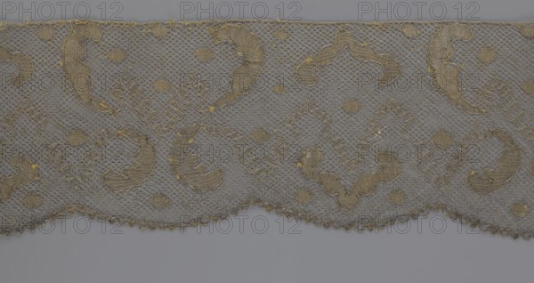 Lace fragment, lace, 18th century, Europe, lace, fabric, width 20 cm, length 120 cm, A strip of delicate lace in shades of gray and gold fills the upper portion of the image, featuring a repeating scalloped edge and a net-like background; the lace has several ornate, curved embroidered motifs and small circular adornments in metallic gold thread, with tiny loose threads and slight fraying visible along some edges; the lower portion shows a smooth gray surface beneath the lace.