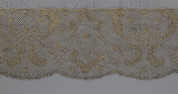 Lace fragment, lace, 18th century, Europe, fabric, width 20 cm, length 120 cm, A piece of lace fabric with a repeating decorative pattern, showing a scalloped lower edge and a straight upper edge, woven in a mesh net structure with embroidered motifs; the lace appears in shades of beige and gold against a gray background, with areas of denser stitching forming curved and circular shapes, small rounded fillers, and ornamental loops, and with visible thread details and fringe along the top edge.
