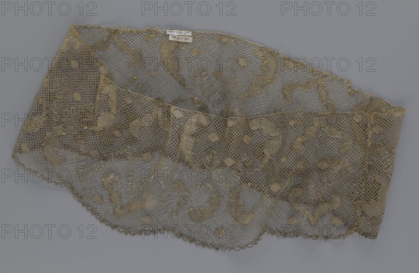 Lace fragment, lace, 18th century, Europe, lace, fabric, width 20 cm, length 120 cm, A delicate rectangular piece of openwork fabric laid flat against a solid background, showing a net-like mesh with ornate embroidered motifs and scalloped edging; the fabric appears sheer and lightweight with a pattern of curved and rounded shapes integrated into the mesh, and a few areas where threads are slightly frayed. The visible colors are shades of muted gold and beige for the fabric and embroidery, set against a uniform cool gray background.