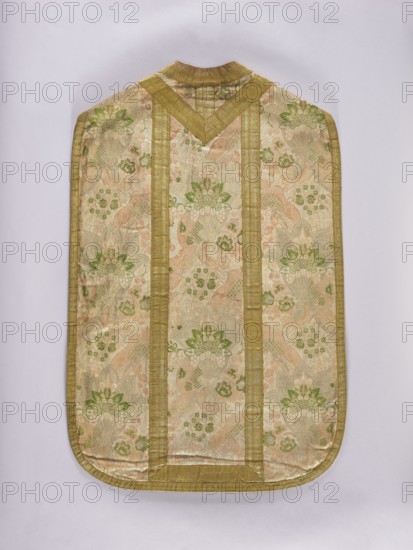 Chasuble, Lace and floral ornaments; pattern of wavy ribbons with fan-shaped bouquets of small flowers surrounded by a serrated collar; background filled with small curling flowers. Columns trimmed with wide gold galloon, edges narrower. Linen lining, pink. 1st quarter of the 18th century, France. Liturgical vestment. Length 101 cm, width 66 cm back., A single ornate textile panel with a rounded bottom and a small neck opening at the top, displayed flat against a pale background. The fabric shows a repeating floral and foliate pattern in muted shades of gold, beige, pale pink, and green, with areas of sheen suggesting metallic threads. Broad vertical bands and a V-shaped yoke of wider gold trim run along the length and near the top, and a narrow gold border edges the entire piece. The floral motifs include stylized blossoms and leafy clusters in green and pink set against a beige-gold ground, with subtle tonal variations and a slightly raised woven texture.