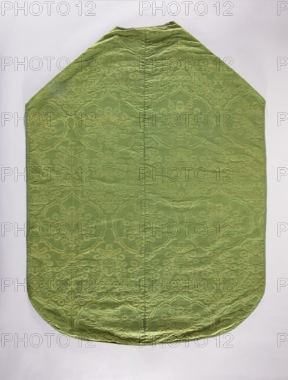 Chasuble;, 16th century, Italy. Liturgical vestment church paraments, length 129 cm, width 97 cm back. Design: plant ornaments and flowers — a seven-leafed rose with a floral thistle flower; above the rose is a small crown and above that the same floral thistle motif. From the crown two intertwined floral branches branch down, creating a roof-like form; this motif repeats in horizontal rows. Trimmed with a narrow green ribbon; green cotton lining. Function: sacred art, liturgical vestment for churches and chapels. Repossession, A single large fabric panel laid flat with a pointed top and rounded bottom, showing a subtle raised floral and scroll damask pattern across its surface; the fabric appears primarily light green with slightly darker green raised motifs and gentle wrinkles and seams running vertically; the background behind the panel is a pale off-white.