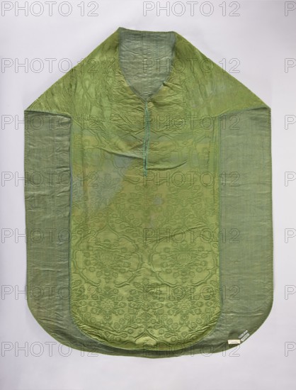 Chasuble, repossession, Design: plant ornaments and flowers—roses and thistle flowers—forming a seven-leafed rose with a floral thistle flower. Above the rose at the top is a small crown, and above that the same floral thistle motif. From this crown two intertwined floral branches branch down, creating a kind of roof; this motif repeats alternately in horizontal rows. Trimmed with a narrow green ribbon. Green cotton lining. 16th century, Italy. Liturgical vestment church paraments/sacred art, length 129 cm, width 97 cm back., A flat garment laid out against a pale background, primarily green with variations from light to darker green; it has a rounded lower edge and a pointed upper section forming a neckline opening, with a narrow strip running down from the neckline. The central panel shows a subtle woven floral and lattice pattern visible as slightly darker green motifs against a lighter green field, while the side panels are a more translucent, plain green fabric. The texture shows ...