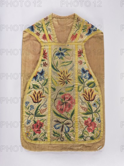 Chasuble, Chasuble made of woolen canvas embroidered with patterns of large, fantastic, diverse flowers on a common stem. Embroidery colors: green, blue, yellow, brown, red. Trimmed with yellow silk braid. Cotton lining. 18th century, Silesia. Liturgical vestment. Length 106 cm, width 65.5 cm., A garment laid flat against a pale background, made of a light tan or beige base fabric with a decorative front panel featuring embroidered floral motifs. The central area and side panels show a range of colorful embroidered flowers and leaves in shades of blue, red, pink, yellow, green, and touches of cream, framed by yellow trim. The embroidery includes stylized blooms with layered petals and curved stems, surrounded by leafy shapes and small rounded floral elements. The shoulder and neckline areas have matching embroidered sections with similar color palette and yellow edging. The overall shape tapers toward the waist and flares slightly toward the hem, with the lower edge following the garme...