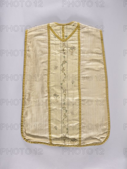 Chasuble, Liturgical vestment made of three types of silk fabrics, trimmed with yellow silk braid; cotton lining green on the front, navy blue on the back. Front features an embroidered column of white silk grosgrain embroidered with silk threads in patterns of small floral sprigs; three sprigs placed sparsely, likely cut from another piece and added. Back column and neckline made of silk weft-weave fabric brocaded with silk yarn and silver thread in a striped pattern with a wavy floral tendril and small floral sprigs. Sides made of cream-colored striped silk fabric. Decorated with flowers, leaves, and plant ornaments. 2nd half of the 18th century. Place of origin: Poland; back: France. Length 98.5 cm, width 68 cm., A flat garment displayed against a pale background, made of a shiny cream fabric with a central vertical panel bordered by narrow golden-yellow trim and decorated with a delicate vertical vine of embroidered small flowers and leaves in muted green and pale brown tones; the ...