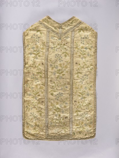 Chasuble, liturgical vestment. White chasuble covered with colorful and silver embroidery in a small floral pattern: forget-me-nots, lilies of the valley, daffodils, grapes, carnations, etc. Trimmed with silver braid. Cotton lining. 2nd half of the 18th century, Poland. Length 100 cm, width 61 cm back., A long, sleeveless textile panel with a V-shaped top, primarily pale cream in color, decorated with a repeating floral pattern in soft green and light blue, with touches of pale yellow; the fabric shows areas of sheen and subtle wrinkling, and is bordered and sectioned by narrow metallic trim in a muted silver tone; the background behind the panel is a smooth, light neutral color.
