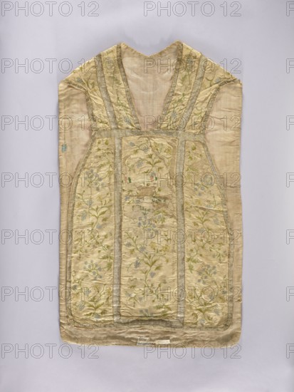 Chasuble, liturgical vestment, 2nd half of the 18th century, Poland. White chasuble covered with colorful and silver embroidery in a small floral pattern forget-me-nots, lilies of the valley, daffodils, grapes, carnations, etc., trimmed with silver braid; cotton lining. Dimensions: length 100 cm, width 61 cm back., A single sleeveless garment laid flat, showing a V-shaped neckline and a straight, rectangular lower body with slightly angled sides; the fabric appears pale beige or cream overall with panels of embroidered floral patterns in muted green and pale blue, outlined and divided by narrow metallic trim in a silvery color; the embroidered designs show winding stems and small blossoms across several vertical panels; the material has a slightly worn and wrinkled texture with visible seams and stitched borders; the background surface is a light neutral color.