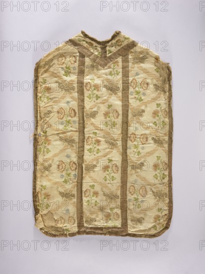 Chasuble, liturgical vestment embroidered on white rep with pastel-colored silks and gold thread in patterns imitating brocade: wavy ribbons and floral bouquets forming a network arrangement. Columns emphasized with gold galloon stitching. Cotton lining. Embroidery colors: green, ochre, beige, blue. 2nd half of the 18th century, Poland. Length 96 cm, width 66 cm back., A worn textile garment laid flat on a pale background showing a light cream base with a floral pattern in shades of green, brown, pale blue, and soft pink, framed by darker brown vertical and edge trim. The fabric has a V-shaped neckline at the top and visible creases, fraying, and small tears along the edges and corners. The floral motifs repeat across the surface with leafy elements and rounded blossoms, and the darker trim forms parallel vertical bands and borders around the piece. The overall colors visible are cream, brown, green, pale blue, and soft pink.