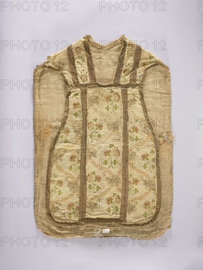 Chasuble, embroidered on white rep with pastel-colored silks and gold thread in patterns imitating brocade: wavy ribbons and floral bouquets forming a network arrangement; columns emphasized with gold galloon stitching; cotton lining; embroidery colors: green, ochre, beige, blue. Liturgical vestment, 2nd half of the 18th century, Poland. Length 96 cm, width 66 cm back., Keywords: liturgical vestments, flowers, plant ornaments, church paraments, sacred art, churches and chapels furnishings., A sleeveless garment laid flat against a pale background, primarily in shades of beige and cream with a patterned central panel featuring floral motifs in green and muted brownish-pink tones; the garment has vertical and curved bands in a darker tan or brown framing the patterned sections, and visible frayed edges and worn areas with some discoloration in orange-brown; the fabric appears textured with a slightly shiny finish in places and a matte backing in others, showing small irregularities and r...