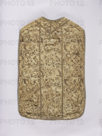 Chasuble., Liturgical vestment, 18th/19th century, France. Embroidered with flowers and plant ornaments; columns highlighted with narrow braid stitching and gold-brown darts. Edges trimmed with gold-silver braid. Blue linen lining. Length 95 cm, width 64 cm back., An ornate textile panel with a rectangular shape and rounded bottom corners, displayed flat against a pale background; the fabric shows an intricate floral and foliate woven pattern in gold and brown tones with subtle highlights of lighter gold, and there is a decorative vertical banding in darker brown that creates panel divisions and a chevron-shaped detail near the top, while the edges are trimmed with a narrow border that appears in contrasting muted green and brown.