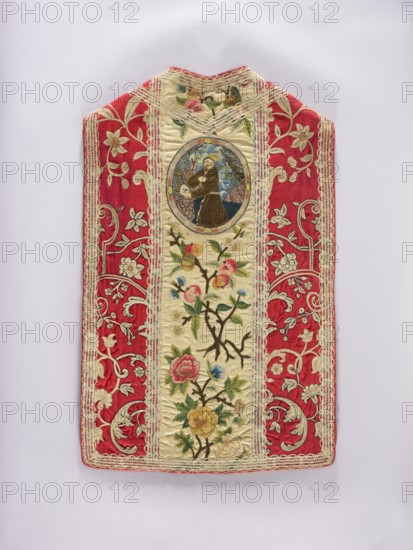 Chasuble, 17th century, Poland. Liturgical vestment, length 95 cm, width 60 cm back. Cotton lining. Trimmed with period white lace. Predominant colors: white for the columns, red for the sides, and yellow, pink, blue, and white for the ornament. Decoration: columns with plant ornaments — at the back, at the top in an oval embroidered kneeling St. Anthony Anthony of Padua, 1195–1231, below branches of apple blossoms and fruit; at the front, branches with apple blossoms and fruit. Sides: ornament with stylized plant twigs. Iconography: Anthony of Padua; function: church paraments / sacred liturgical vestment., An ornate textile panel with a central cream-colored vertical band framed by a decorative lace trim, flanked by wide red panels patterned with pale floral and scroll embroidery; the cream band features embroidered flowering branches with pink, yellow, blue, and green floral motifs and a circular embroidered medallion near the top showing a figure in brown robes against a blue and t...