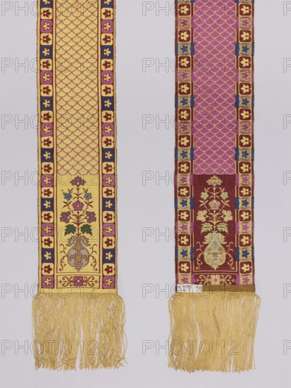Half-belt. Double-sided silk half-belt. Each head features a single motif of a flower bouquet in a vase. Center with a carp scale motif. Vertical borders divided into small quarters, each with leaf motifs; the lower horizontal borders upper ones missing contain single rosettes. Borders in the form of straight lines. Background colors: yellow and brown; ornament colors: blue, purple, yellow, green, silver. Fringe sewn on; materials: silk, straw. Weave density approx. 18/25 x2. Date: 1890–1910. Function: clothing accessory, national costume; length 345 cm; width 12 cm. Polish kontusz/double-sided belt style; men's clothing accessory. Plant ornaments, rosette, carp scale ornament., Two long vertical woven bands hang side by side against a pale background, each ending in a fringe of golden yellow threads. The left band features a golden yellow field with an allover scalloped pattern in a darker tone and a narrow border of small floral motifs in pink, blue, and maroon; near its lower sectio...