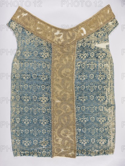 Chasuble, liturgical vestment. Genoese velvet, patterned, convex, with a regularly repeating motif of a stylized bouquet of flowers in vases enclosed in a thick heart-shaped net; mesh fastened with palmettes. Colors: sapphire blue ornament on a cream-gold background; columns of solid material in gold-beige. Ornamentation partly illegible due to large defects; made of stylized plant forms. 15th/16th century. Place of origin possibly Italy or France; sides: Genoa. Dimensions: length 122 cm, width 64 cm back., A rectangular textile panel with a central vertical strip of patterned tan fabric flanked by wider panels of patterned blue fabric, the blue panels showing a repeating ornate motif in lighter blue and tan tones; the tan strip has a swirling floral-like pattern in slightly darker tan and is bordered by narrow decorative seams; the top edge widens into a shallow V-shaped yoke of the same tan fabric with visible worn patches and lighter areas; the blue side panels exhibit areas of wear...