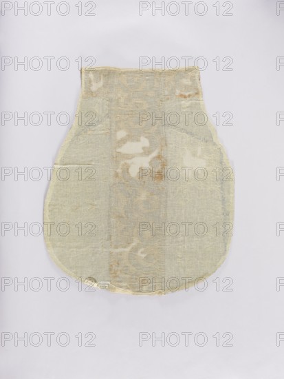 Chasuble, liturgical vestment. Genoese velvet chasuble, patterned, convex, with a regularly repeating motif of a stylized bouquet of flowers in vases enclosed in a thick heart-shaped net. Mesh fastened with palmettes. Colors: sapphire blue ornament on cream-gold background; columns of solid material in gold-beige. Ornamentation partially illegible due to large defects; composed of stylized plant forms. 15th/16th century, Italy possibly France or Genoa. Length 122 cm, width 64 cm back., A single fabric piece with a rounded bottom and a narrower top is laid flat against a pale background; the fabric shows a worn, slightly translucent texture with uneven areas and frayed edges, and displays a muted palette of beige, off-white, light tan, and faint grayish tones with subtle mottled patches of darker tan.
