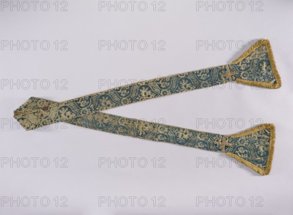 Stole, church paraments; embroidered with flowers, leaves, and crosses; liturgical vestment with plant ornaments; sacred art; for churches and chapels furnishings; widened trapezoidal ends trimmed with a short yellow cotton fringe; lined with silk fabric; 15th century, Genoa, Italy; height 236 cm, width 21 cm., A long decorative fabric band laid out on a pale background, with a pair of tapered ends each finished with triangular flared sections trimmed in fringe; the fabric shows a repeating ornate pattern in teal and beige with areas of wear and faded color, and the fringe and some edging are a golden yellow; the band crosses near one end and has a small sewn patch visible where layers overlap.