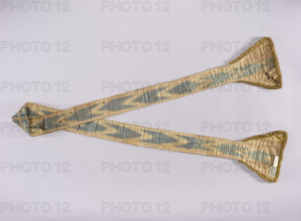 Stole, church paraments; features flowers, leaves, crosses; liturgical vestment with plant ornaments and sacred art; churches and chapels furnishings. Widened trapezoidal ends trimmed with a short yellow cotton fringe. Lined with silk fabric. 15th century, Genoa, Italy. Height 236 cm; width 21 cm., A long, narrow fabric sash laid out in an overlapping X shape on a pale background, showing a silky texture with worn areas and small holes; the sash has a faded pattern of bluish-gray streaks on a golden-beige ground, with triangular widened ends edged in golden fringe and one end showing a small patch of blue and cream patterned fabric.