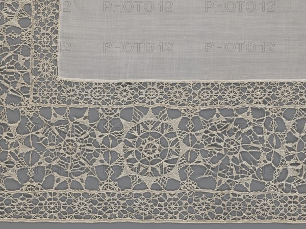 Doily, geometric ornaments, rosettes ornament, doilies. Smooth center. Edge made of white linen lace with rosette motifs: large rosettes in the center and rows of small rosettes on the edges. Punto in tela lace, ca. 1600, Italy. Lace, fabric. Height 79 cm, width 70 cm., The image shows a close-up of a textile with a plain rectangular inner fabric in a light beige color bordered by an intricate openwork lace edging in a slightly darker beige tone against a medium gray background; the lace features a variety of geometric and floral-inspired motifs connected by delicate thread bridges, creating circular and star-like patterns and irregular open spaces, with visible fine woven texture on the central fabric and a scalloped outer edge of the lace.