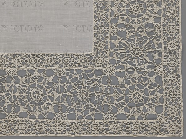 Doily, geometric ornaments, rosettes ornament, doilies. Smooth center. Edge made of white linen lace with rosette motifs: large rosettes in the center and rows of small rosettes on the edges, punto in tela lace, ca. 1600, Italy. Lace, fabric; height 79 cm, width 70 cm., The image shows a section of lace fabric with a solid woven square of cloth in the upper left corner bordered by an intricate openwork lace design. The lace is composed of a network of delicate loops and starburst-like motifs, forming geometric and floral patterns connected by fine threads. The visible colors are shades of off-white or cream for the fabric and lace, contrasted against a muted gray background that shows through the open spaces. The textures range from the smooth, tightly woven cloth to the raised, airy, and filigree-like texture of the lace.