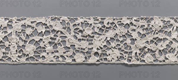 Lace fragment, needle lace, floral ornaments with a pattern of large, thick branches with stylized flowers and leaves; details executed in relief; joints rarely with rosettes snowflakes. 2nd half of the 19th century, Italy. Materials: lace, fabric. Dimensions: width 15 cm, length 248 cm. Creator, purchase provenance., A wide horizontal strip of openwork lace with an intricate floral and foliate pattern rests against a neutral background; the lace appears ivory or off-white and features interconnected motifs including stylized flowers, leaves, scrolls, and tendrils formed by solid embroidered areas and delicate connecting bars, with a narrow decorative border along the lower edge. The background visible through the lace is a smooth mid-gray, and subtle shadows from the raised embroidery create gentle tonal variations where the lace overlaps the surface. The overall impression shows the contrast between the pale lace and the gray background, highlighting the texture and three-dimensional...
