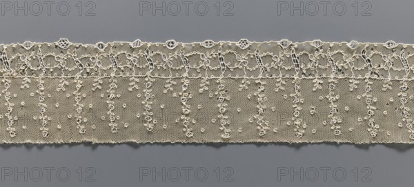 Fragment of lace, lace, ornament: continuous vertical twigs at the top with points between them; at the bottom a strip of edge ornament where plant and geometric tendrils intertwine. Late 18th century, possibly Germany or France. Fabric lace, width 9.5 cm, length 67 cm., A horizontal strip of lace in an off-white or cream color stretches across a gray background, featuring a scalloped top edge with a row of small circular openings and an ornate looping embroidered border beneath; below that are several vertical braided or beaded-looking embroidered columns ending in tiny leaf-like motifs, set against a fine netted ground sprinkled with a few small raised circular dots and delicate embroidered clusters, with the lower edge of the lace slightly scalloped and uneven.