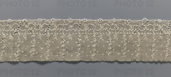 Fragment of lace, lace, ornament: continuous, vertical twigs at the top with points between them, at the bottom a strip of edge ornament where plant and geometric tendrils intertwine. Late 18th century, possibly Germany or France. Material: lace fabric; width 9.5 cm, length 67 cm., A horizontal strip of delicate lace with a scalloped top edge and a straight bottom edge lies against a plain background; the lace is a light beige or off-white color with an intricate openwork pattern including a decorative upper border of looping floral-like motifs and a lower field of a fine hexagonal mesh scattered with small raised knot-like dots and vertical series of tiny open rings; the background visible through the lace is a smooth medium gray, and shadows emphasize the texture and openness of the fabric.
