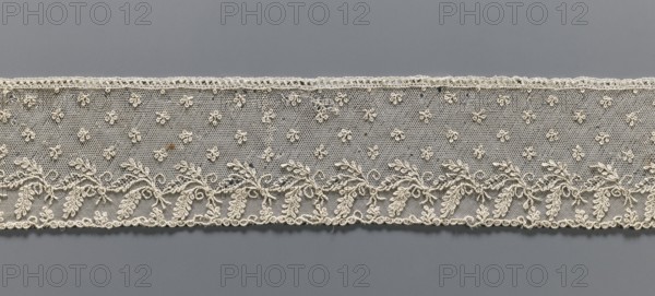 Fragment of lace, Alençon lace, decorated with small twigs in projections and a tendril with feathery leaves running parallel to the edge against a mesh background, 2nd half of the 18th century, France, lace, width 8.5 cm, length 54 cm, A horizontal strip of delicate lace in an off-white or cream color stretches across a neutral gray background; the lace features a fine hexagonal mesh filled with a scattered pattern of small rosette-like motifs and a continuous embroidered border along the lower edge composed of sinuous vine shapes with leafy clusters and curled tendrils, while the top edge has a narrow scalloped trim; the overall texture appears intricate and slightly raised against the smooth gray surface.