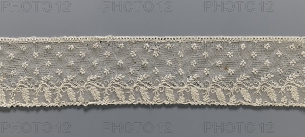 Fragment of lace, Alençon lace decorated with small twigs in projections and a tendril with feathery leaves running parallel to the edge against a mesh background. 2nd half of the 18th century, France . Lace, width 8.5 cm, length 54 cm., A horizontal strip of delicate lace in an off-white or cream color is set against a neutral gray background; the lace features a fine hexagonal mesh with a pattern of small raised floral motifs scattered across the upper area and a denser embroidered border along the lower edge that shows swirling leafy and vine-like shapes, scalloped trim, and tiny loops; the overall texture appears slightly uneven with occasional thicker threads and small irregularities in the lacework.