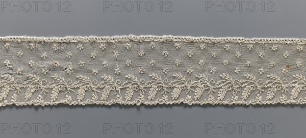 Fragment of lace, Alençon lace, decorated with small twigs in projections and a tendril with feathery leaves running parallel to the edge against a mesh background. 2nd half of the 18th century, France, lace, width 8.5 cm, length 58.5 cm., A horizontal strip of delicate lace in off-white and cream tones stretches across a gray background, featuring a fine netted field with a pattern of small floral motifs and a lower border of intertwined leafy garlands and scrolls, with scalloped edges and a few small areas of darker discoloration.