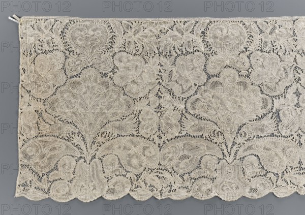 Lace fragment, unknown creator; purchase provenance. Brussels lace, early 18th century. Pattern with motifs of a large leafy-palmette bouquet emerging from a vase similar to a pear; above the bouquet is a heart-shaped palmette. Few de brides-style connections with picots. Lace is dense and likely of the opaque type. Lower edge gently serrated, upper edge straight. Materials/technique: lace. Width 41 cm, length 356 cm., A large panel of intricate lacework in off-white and cream tones lies flat against a gray background, showing a repeating floral and foliate pattern with scalloped edges. The lace features a few prominent stylized blossoms and leaves formed by denser embroidered areas surrounded by openwork, with several looping and curving motifs connecting the elements. The texture appears delicate and slightly raised in places where the threads are more tightly worked, and the scalloped hem showcases rounded, petal-like shapes. The overall colors visible are off-white, cream, and gray...