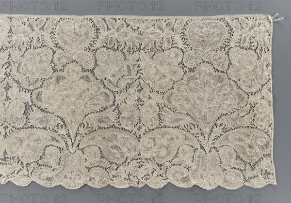 Lace fragment, Brussels, early 18th century. Pattern with motifs of a large leafy-palmette bouquet emerging from a vase similar to a pear. Above the bouquet is a heart-shaped palmette. Few de brides-style connections with picots. The lace is dense and therefore probably of the opaque type. The lower edge is gently serrated, the upper one straight. Brussels lace; materials: lace. Width 41 cm, length 356 cm., A large rectangular piece of intricately patterned lace laid flat, showing an overall cream color with subtle variations in tone and a gray background visible through many open areas; the lace features a repeating floral and foliate motif with ornate curved shapes, scalloped lower edge, and a few small irregular holes within the mesh; the texture appears delicate and finely worked with dense embroidered outlines and more open netted sections, and there is a narrow straight upper edge with a slightly different finish.