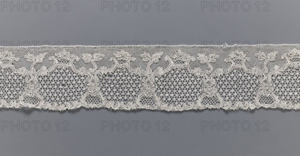 Lace fragment, purchased provenance noted, lace, Malines Mechelen lace, shell and plant pattern; mesh background: point de neige and drochelle; 2nd half of the 18th century; Mechelen; lace; width 7.7 cm, length 521 cm, A horizontal strip of delicate lace laid against a smooth background, showing an intricate repeating floral and scrollwork pattern with a scalloped lower edge and a straight upper edge; the lace has open hexagonal mesh sections interspersed with denser embroidered motifs that form stylized flowers and curving vines, and the piece appears in shades of off-white and cream against a medium gray backdrop.