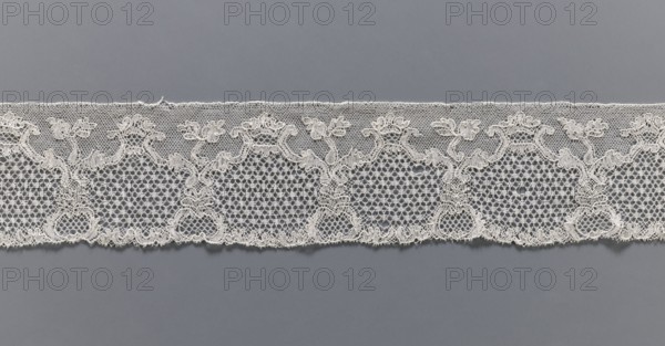 Lace fragment, purchased provenance noted, lace, Malines Mechelen lace, shell and plant pattern; mesh background: point de neige and drochelle; 2nd half of the 18th century; Mechelen place of origin; lace; width 7.7 cm, length 521 cm, A horizontal strip of delicate lace laid flat against a plain background, showing a repeating ornate pattern with floral and scroll-like motifs connected by fine netting; the lace appears off-white or cream with intricate openwork and scalloped lower edge, and the background is a uniform medium gray.