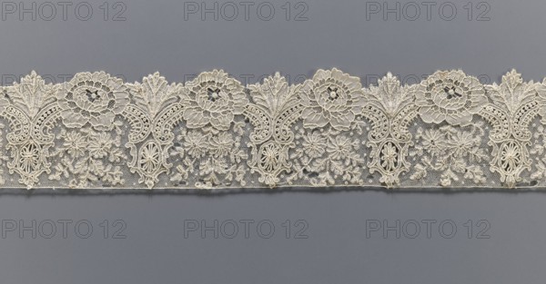 Lace fragment, purchase provenance noted; lace with plant ornaments. Serrated on one side, with a pattern of flowers, fan-shaped leaves, and ovoid rosettes above them. Rose flowers between the leaves, and sprigs of other small flowers above them. Background fond de gaze. 2nd half of the 19th century, Brussels; lace, width 8.5 cm, length 121 cm., A horizontal strip of intricately patterned lace with a scalloped top edge rests against a solid background; the lace is a creamy off-white color with floral and leaf motifs formed by raised threadwork and open netting, showing repeating rosette shapes, stylized leaves, and clusters of small embroidered flowers, while the background is a smooth muted gray.
