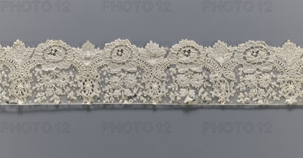 Lace fragment, lace with plant ornaments. Serrated on one side, with a pattern of flowers, fan-shaped leaves, and ovoid rosettes above them. Rose flowers between the leaves, and sprigs of other small flowers above them. Background fond de gaze. 2nd half of the 19th century, Brussels, lace, width 8.5 cm, length 121 cm, A horizontal strip of ornate lace with an intricate floral and scrollwork pattern in off-white and cream tones set against a solid gray background; the lace features a scalloped upper edge with repeating circular flower motifs and leaf-like arches, a lower straight edge, a mesh-like netting infill, and a few raised embroidered floral clusters and decorative oval medallions, all rendered in pale ivory shades with subtle shadows and highlights.
