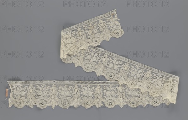 Lace fragment, lace with plant ornaments. Serrated on one side, with a pattern of flowers, fan-shaped leaves, and ovoid rosettes above them. Rose flowers between the leaves, and sprigs of other small flowers above them. Background fond de gaze. 2nd half of the 19th century, Brussels; lace, width 8.5 cm, length 127 cm, A length of delicate lace ribbon with an intricate floral and scroll pattern arranged in a sinuous shape against a flat background; the lace is a pale cream color featuring scalloped edges and repeating ornate motifs, and the background is a medium gray.