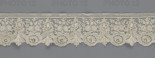 Lace fragment, lace with plant ornaments. Serrated on one side, with a pattern of flowers, fan-shaped leaves, and ovoid rosettes above them. Rose flowers between the leaves, and sprigs of other small flowers above them. Background fond de gaze. 2nd half of the 19th century, Brussels; lace; width 8.5 cm, length 127 cm., A horizontal band of delicate lacework with an ornate repeating floral and scroll pattern, displayed against a muted gray background. The lace is a pale cream color and features a scalloped lower edge with a few large circular floral motifs, several fan-shaped leaf elements, and a group of smaller flowers and leaves woven into a fine net ground. The upper edge has a narrow openwork border with a rectangular mesh. The overall texture appears intricate and raised in places, showing loops, cords, and denser embroidered areas contrasting with the sheer net. The visible colors are cream and gray.