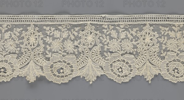 Lace fragment, purchase provenance; lace with plant ornaments. Serrated on one side, pattern of flowers, fan-shaped leaves, and ovoid rosettes above them. Rose flowers between the leaves, and sprigs of other small flowers above them. Background fond de gaze. 2nd half of the 19th century, Brussels; lace, width 8.5 cm, length 127 cm, A horizontal strip of delicate lace in off-white spans the image against a gray background, featuring an intricate repeating pattern of floral motifs and ornate scrollwork; a few large scalloped floral rosettes sit along the lower edge, connected by ornate fan-shaped and curved designs with openwork details, while a group of smaller embroidered flowers and leafy sprigs fill the central netted ground above them; the top edge shows a narrow band of more geometric, ladder-like lacework; overall colors visible are off-white lace and a medium gray background.