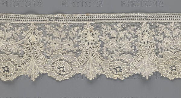 Lace fragment;, purchased provenance noted; lace with plant ornaments. Serrated on one side, with a pattern of flowers, fan-shaped leaves, and ovoid rosettes above them. Rose flowers between the leaves, and sprigs of other small flowers above them. Background fond de gaze. 2nd half of the 19th century, Brussels. Lace; width 8.5 cm, length 127 cm., A horizontal strip of delicate lace in off-white and cream tones stretches across a muted gray background, featuring an intricate repeating floral and ornamental pattern with scalloped lower edge; the lace shows a mesh-like netting filled with embroidered flowers, leaves, and ornamental medallions, with several larger floral rosettes and a few ornate vertical motifs that rise from the scallops, connected by swirling embroidered lines and tiny circular details, and the top edge has a narrow woven band in the same cream color.