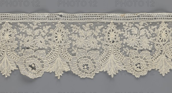 Lace fragment, lace with plant ornaments. Serrated on one side, with a pattern of flowers, fan-shaped leaves, and ovoid rosettes above them. Rose flowers between the leaves, and sprigs of other small flowers above them. Background fond de gaze. 2nd half of the 19th century, Brussels, lace, width 8.5 cm, length 127 cm, A length of delicate lace with an intricate floral and scroll pattern placed against a solid background; the lace shows a scalloped lower edge with large stylized flower motifs and fan-like tassel shapes, a middle section of open netting filled with embroidered blossoms and leaf sprigs, and an upper border of a tighter woven band; visible colors include off-white or cream for the lace and a gray background.