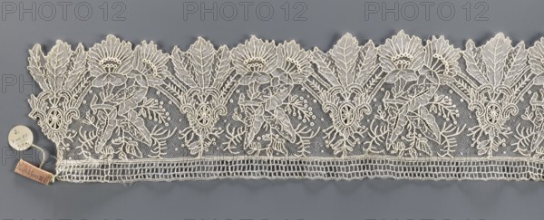 Lace fragment, Plant ornaments; with a pattern of stylized leaves and flowers, serrated on one side. Background fond de gaze. 2nd half of the 19th century, Brussels. Lace, width 11 cm, length 147 cm., A horizontal strip of delicate lace with an intricate floral and leaf pattern in off-white set against a gray background, showing a scalloped top edge formed by large stylized leaves and flower shapes, a central area of fine netting filled with embroidered botanical motifs including curved branches and clustered beadlike accents, and a lower straight edge featuring a rectangular openwork band; a small round tag and a small rectangular fabric tag are attached near one end, and visible colors include off-white, gray, and a small patch of pale tan on the rectangular tag.
