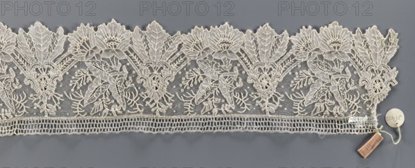 Lace fragment, Plant ornaments; pattern of stylized leaves and flowers, serrated on one side. Background fond de gaze. 2nd half of the 19th century, Brussels. Lace; width 11 cm, length 147 cm., A horizontal strip of lace with an intricate floral and leafy pattern laid flat on a gray background; the lace is off-white or cream in color and features repeating scalloped upper edges with large stylized flowers and pointed leaves, detailed openwork netting filling spaces between motifs, and denser embroidered outlines that create textured petals and foliage. Along the lower edge the lace has a band of rectangular openwork and a narrow embroidered border. A small round tag and a rectangular cloth tag attached by a thin string rest on the gray surface near one end of the lace; the visible colors are cream, gray, and a small area of muted brown on the rectangular tag.