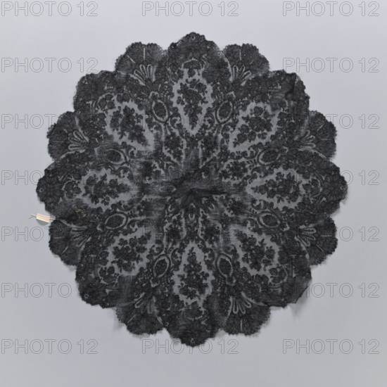 A fragment of lace umbrella cover, lace; patterns arranged radially from a central rosette; outer edge serrated with alternating fan-shaped leaves and large flower rosettes; above the rosette, leaves and C shapes frame an oval field containing a branch with flowers and rosebuds; 19th century, France; diameter 60 cm., A circular lace textile with a scalloped edge displayed flat on a light gray background, featuring an intricate floral and paisley-like pattern radiating from the center; the lace appears predominantly black with varying densities creating lighter gray areas where the background shows through, and there is a small pale rectangular tag attached near the outer edge.